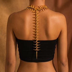 May include: A gold-toned metal body chain featuring a spine-like design with sharp, pointed segments that extend from the neck down the center of the back, resting over a plain black strapless top. The piece has a metallic finish and structure.