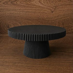 Könnte beinhalten: Ein mattschwarzer Tortenständer mit einer runden Platte, die einen vertikal gerippten Rand aufweist, und einem passenden, sich verjüngenden, kannelierten Sockel. Das Design besteht aus einer flachen, glatten Oberfläche auf einem stabilen Fuß.