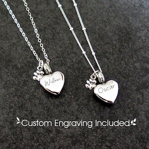 Puede incluir: Dos collares plateados con colgantes en forma de corazón y dijes de huellas de pata. Uno está grabado con "Willow", el otro con "Oscar". La imagen incluye el texto "Custom Engraving Included."