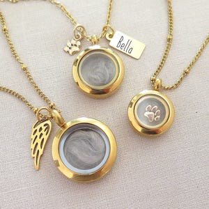 May include: Three gold-colored memorial necklaces with round lockets. One locket has a paw print, another has a small tag that says "Bella", and the third has a wing charm. The lockets contain a gray substance and hang on delicate chains.