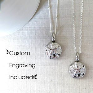 Puede incluir: Dos collares de plata con colgantes de dólar de arena. El texto "Grabado personalizado incluido" es visible debajo de los collares.
