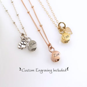 Puede incluir: Tres collares en plata, oro rosa y oro con dijes grabados personalizados. Los dijes son redondos y llevan los nombres "Zoe", "Chloe" y "Finn". El collar de plata también tiene un pequeño dije en forma de huella de pata.  "Grabado personalizado incluido" está escrito debajo de los collares.