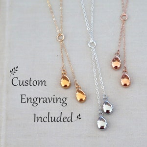 Könnte beinhalten: Drei Halsketten im Lariat-Stil in Gold-, Silber- und Roségoldtönen, jeweils mit einer feinen Kette und zwei tropfenförmigen Anhängern. Der Text "Custom Engraving Included" ist links mit kleinen Blattsymbolen aufgedruckt.