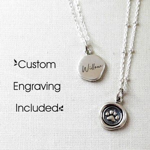 May include: Two silver necklaces with delicate chains. One pendant is a smooth round disc engraved with the name Willow. The other pendant features a raised paw print design. The text Custom Engraving Included is visible on the left side of the image.