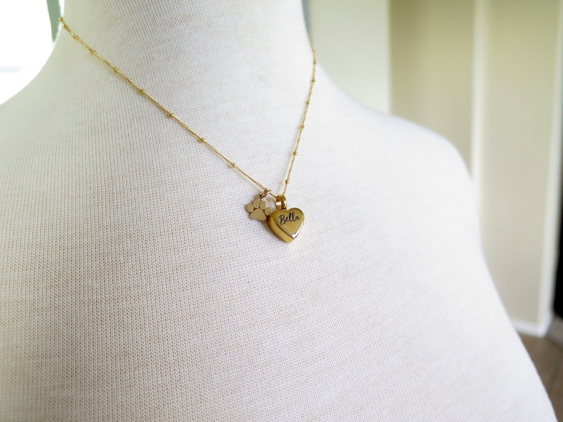 May include: Gold necklace featuring a heart-shaped pendant engraved with the name "Bella" and a small paw print charm.