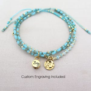 May include: A layered bracelet with turquoise and gold beads. Two gold charms hang from the bracelet, one with the name "Zoey" engraved. The text "Custom Engraving Included" is below the bracelet.