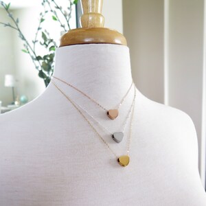 May include: Three layered necklaces with heart-shaped pendants. The top necklace has a rose gold heart, the middle necklace has a silver heart, and the bottom necklace has a gold heart.