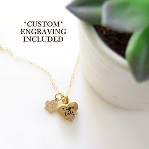 May include: Gold necklace featuring a heart-shaped pendant and a paw print charm. The heart pendant is engraved with the names "Tiger Lily". The necklace is displayed on a white surface with a potted plant in the background. *CUSTOM ENGRAVING INCLUDED*