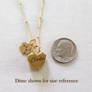 May include: Gold necklace with a heart charm engraved with the name "Charlie" and a small paw print charm. A ten pence coin is shown for size reference.