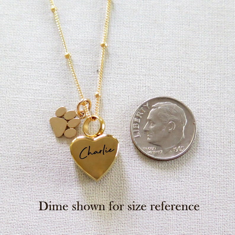 May include: Gold necklace with a heart charm engraved with the name "Charlie" and a small paw print charm. A ten pence coin is shown for size reference.