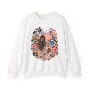 Puede incluir: Una sudadera blanca de manga larga con un estampado gráfico de una persona sentada en una playa al atardecer. El diseño incluye palmeras, flores tropicales, un letrero de madera con texto, un libro, una cámara y fotos polaroid de la costa.