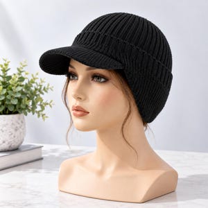 May include: A solid black knit cap featuring a structured brim and a ribbed texture. The hat is designed with a rounded crown and covers the ears, providing a fitted silhouette. The material appears to be a thick, warm yarn construction.