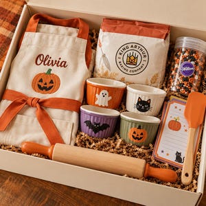 May include: A gift box containing a cream-coloured apron with the name Olivia and a pumpkin embroidered on it, a bag of King Arthur Baking Company flour, a jar of Wilton sprinkles, four small ceramic ramekins with Halloween motifs, a wooden rolling pin with orange handles, a silicone spatula, and a set of illustrated recipe cards.
