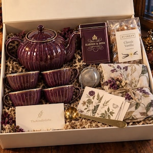 Könnte beinhalten: Eine Geschenkbox mit einer tiefvioletten, gerippten Teekanne und vier passenden Teetassen mit Goldrand, einer Harney & Sons Teedose, einer Tüte McCrea's Classic Vanilla Karamellen, einem Tee-Ei, einem geblümten Tuch, einem goldenen Löffel und einer Karte.