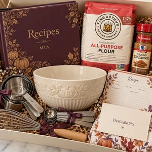 May include: A baking gift set containing a dark purple recipe book labelled Recipes MIA, a 2.27 kg bag of King Arthur all-purpose flour, a bottle of McCormick ground cinnamon, a cream-coloured ceramic bowl, stainless steel measuring cups and spoons, a whisk, a recipe card, and a floral patterned kitchen towel.