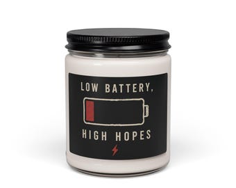 May include: A white candle in a clear glass jar with a black lid. The label is black with the text "LOW BATTERY, HIGH HOPES" in white, featuring a graphic of a battery icon with a red segment and a small red lightning bolt symbol below the text.