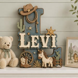 May include: A wooden letter L sign featuring a cowboy theme with a brown hat, lasso, cactus, sheriff star, wagon wheel, and boots. The name LEVI is displayed in large cream-coloured letters. A small horse figure stands near a wooden fence.