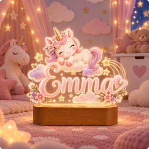 May include: A glowing acrylic night light featuring a cartoon unicorn sleeping on a cloud above the name Emma in pink script. The design includes pastel flowers, stars, and clouds, all mounted on a smooth, oval-shaped wooden base with warm illumination.