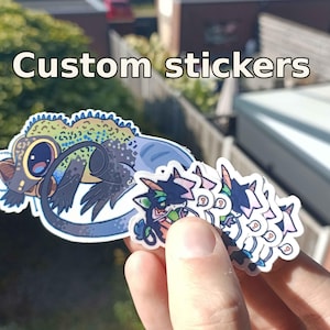 Op de afbeelding: Een verzameling aangepaste stickers met een grote illustratie van een groene en blauwe hagedis met een lange staart, naast verschillende kleinere stickers met een gestileerd personage met roze en blauwe accenten en de tekst Custom stickers.
