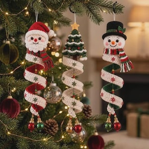 May include: Three hanging crochet ornaments featuring a Santa Claus, a Christmas tree, and a snowman. Each figure has a long, spiraling tail made of cream-colored yarn with red and green snowflake embroidery, finished with small metallic baubles.