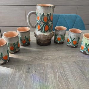 Peut inclure: Une carafe en céramique et six gobelets assortis présentant une base de couleur crème avec un motif répétitif de formes circulaires orange encadrées par des lignes courbes vert foncé. L'ensemble a une finition brune texturée à la base.