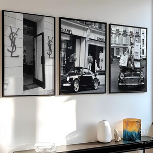 Può includere: Tre stampe da parete incorniciate in bianco e nero. La stampa a sinistra mostra una vetrina Saint Laurent con il logo YSL. La stampa centrale presenta un'auto sportiva nera parcheggiata davanti a una boutique Chanel. Quella a destra mostra una persona.