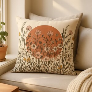 May include: A square throw pillow with a cream-colored background featuring a large, solid terracotta-colored circle in the center. The pillow is decorated with a detailed botanical illustration of various wildflowers and stems in shades of green.