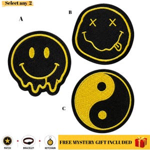 Puede incluir: Tres parches bordados circulares con diseños en negro y amarillo. El parche A es una cara sonriente derritiéndose, el B una cara sonriente con ojos en X y lengua, y el C un símbolo yin-yang. El texto Select any 2 y FREE MYSTERY GIFT INCLUDED es visible.