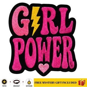 Può includere: Una toppa ricamata con la scritta GIRL POWER in filo rosa acceso su sfondo nero. Un fulmine giallo sostituisce la lettera I e un piccolo cuore rosa chiaro è posizionato sotto la parola POWER. La toppa presenta un bordo nero.