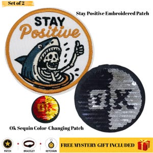 May include: A set of two circular patches. One is an embroidered patch featuring a skeleton shark giving a thumbs up with the text STAY Positive. The second is a round sequin patch that changes color to display the letters OK in silver and black.