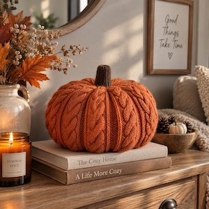 May include: A round, orange, cable-knit fabric pumpkin with a dark brown ribbed stem. The pumpkin sits on top of two stacked books titled The Cozy Home and A Life More Cozy, positioned next to a glass candle jar labeled Pumpkin Spice.
