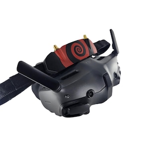 Könnte beinhalten: Ein schwarzes DJI FPV-Headset mit matter Oberfläche, integrierten Antennen und einem schwarzen Stoffkopfband. Der obere Teil zeigt ein rotes und schwarzes Spiralgrafikdesign mit zwei goldenen Antennenanschlüssen, die aus der Oberfläche ragen.