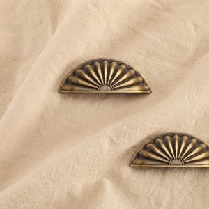 May include: Two semi-circular drawer pulls crafted from antique brass-toned metal. Each pull features a radiating fan-like pattern with deep, curved grooves extending from a central point at the flat base to the rounded outer edge.
