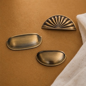 May include: Three brass-toned metal drawer pulls with a brushed finish. One pull is semi-circular with a radiating fan pattern, while the other two are smooth, elongated cup-style pulls of different sizes, all featuring a metallic sheen.