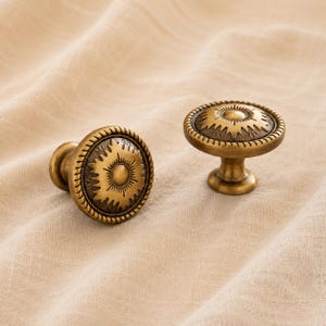 Op de afbeelding: Twee ronde kastknoppen van antiek messingkleurig metaal. Elke knop heeft een centrale cirkelvormige koepel omringd door een zonnestraalpatroon en een touw-getextureerde rand. De knoppen hebben een voetstuk en een gladde, verweerde metalen afwerking.