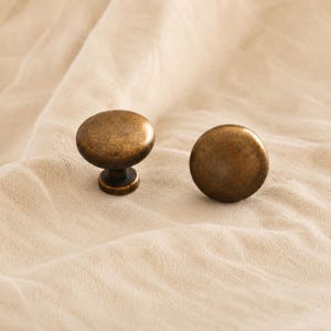 Op de afbeelding: Twee ronde kastknoppen met een verweerde antiek messing afwerking. Elke knop heeft een glad, licht gewelfd oppervlak en een korte cilindrische voet, met een verweerde metalen textuur met donkerbruine en gouden accenten.