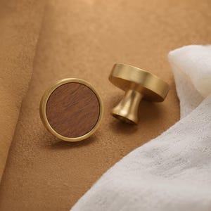 May include: Two circular cabinet knobs featuring a brushed gold metal frame and a flat, circular dark brown wood grain inlay. One knob is positioned face-up, while the other is turned to show its side profile and tapered base.