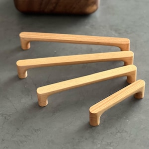 Könnte beinhalten: Vier helle Holzgriffe für Schränke, die in einer diagonalen Reihe von der längsten zur kürzesten angeordnet sind. Jeder Griff hat ein glattes, rechteckiges Stangendesign mit abgerundeten Kanten und zwei zylindrischen Befestigungspunkten.