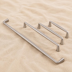 Op de afbeelding: Een set van drie zilverkleurige metalen meubelgrepen in verschillende lengtes. Elke handgreep heeft een glad, cilindrisch ontwerp met licht uitlopende, conische montagepunten aan beide uiteinden voor een strak en uniform uiterlijk.