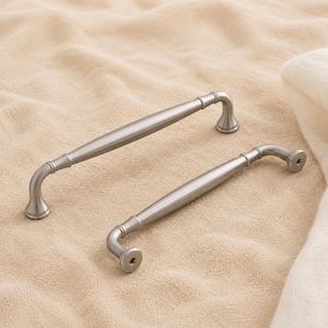May include: Two brushed nickel cabinet pulls with a smooth, tapered cylindrical bar design and rounded mounting bases. Each handle features a subtle curved transition from the central grip to the circular ends, providing a clean, metallic finish.