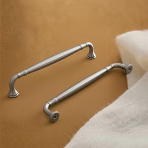 May include: Two brushed silver metal cabinet pulls with a smooth, tapered center bar and curved ends. Each handle features a rounded base at the mounting points, showcasing a clean, metallic finish and a simple, elongated design.
