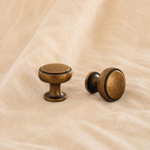 May include: Two round cabinet knobs with a distressed antique brass finish. Each knob features a circular top with a raised centre and a dark recessed ring detail, supported by a tapered base that flares out at the bottom for mounting.