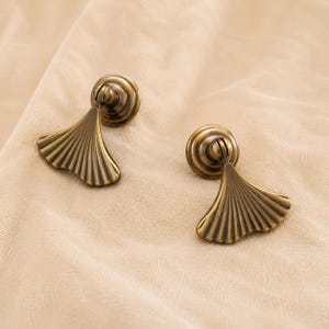May include: Two antique brass drawer pulls featuring a fan-shaped design with vertical ridges. Each pull has a circular mounting base and a hinged, drop-style handle that extends outward, showcasing a metallic finish with dark, recessed detailing.