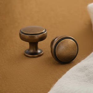 May include: Two round cabinet knobs with a distressed antique brass finish. Each knob features a smooth, slightly domed top surface and a dark, recessed circular ring detail around the edge, set on a tapered base with a dark metal finish.