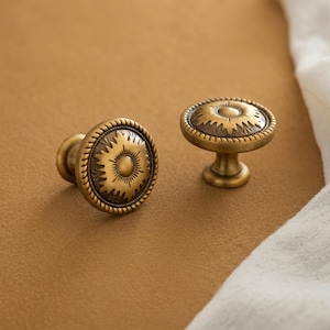 May include: Two round cabinet knobs made of antique brass-toned metal. Each knob features a central circular dome surrounded by a sunburst pattern and a rope-textured border. The knobs have a short, tapered stem and a flat circular base.