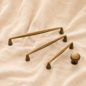 以下が含まれることがあります： 異なる長さのアンティーク真鍮製キャビネットプル3点と、お揃いの丸型キャビネットノブ1点。各パーツは、使い込まれた金属仕上げの先細りでスリムなデザインが特徴で、ハンドルの端には円錐形の取り付けポイントが付いています。