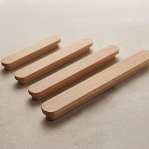 Puede incluir: Cuatro tiradores de armario de madera de color marrón claro dispuestos en una fila diagonal. Cada tirador presenta una forma rectangular alargada y lisa con extremos redondeados y una superficie superior plana, mostrando una veta natural.