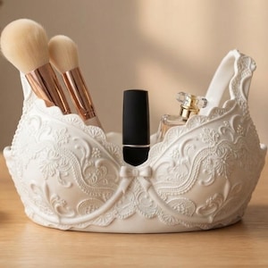 May include: A white, moulded resin organiser shaped like a brassiere, featuring intricate embossed floral and lace-like patterns. It holds two makeup brushes with rose gold ferrules, a black lipstick tube, and a small clear glass perfume bottle.