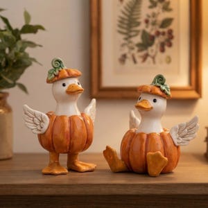 May include: Two ceramic duck figurines, each featuring a white duck body emerging from an orange pumpkin base. Both ducks wear a small orange pumpkin cap with green leaves on their heads and have white feathered wings extended to their sides.
