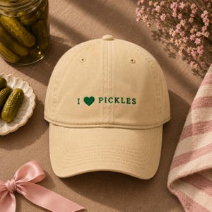 Op de afbeelding: Een beige honkbalpet met de geborduurde groene tekst I heart PICKLES, waarbij het hart een effen groene vorm is. De pet heeft een gestructureerde kroon, een gebogen klep en gestikte oogjes op de bovenste panelen voor ventilatie en stevigheid.